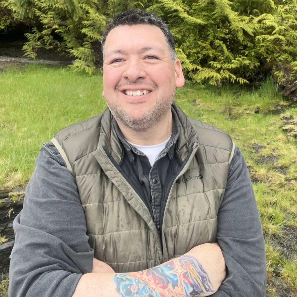 Smiling man wearing a green quilted jacket posing outdoors with lush green foliage behind him.