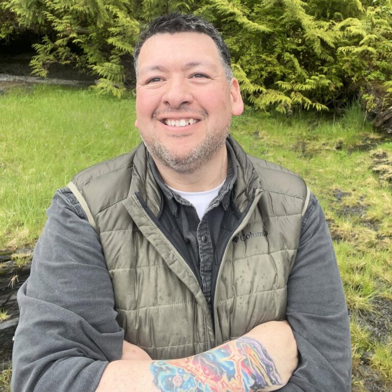 Smiling man wearing a green quilted jacket posing outdoors with lush green foliage behind him.