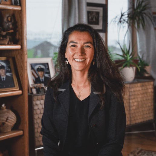 Portrait of a woman with long dark hair, wearing a black blazer, smiling in a cozy living room with photos on a shelf and potted plants in the background.