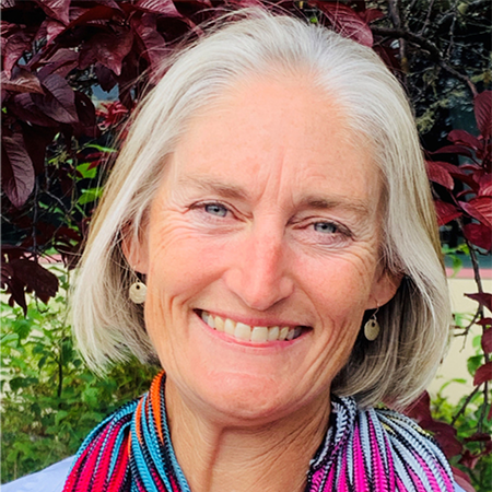 Smiling older woman with silver hair wearing a colorful scarf outdoors, leafy background behind her.