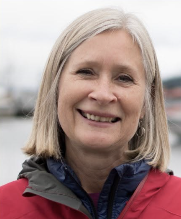 Smiling woman with shoulder-length gray hair wearing a red jacket, outdoors near a body of water.