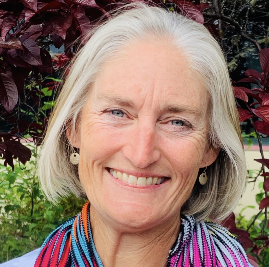 Close-up of a smiling older woman with silver hair, wearing a colorful striped scarf, outdoors among red leaves.
