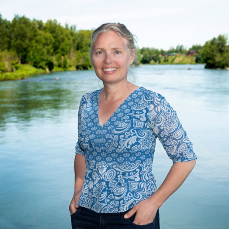 Smiling woman with blonde hair in a blue paisley-patterned top, standing by a calm river with trees in the background.
