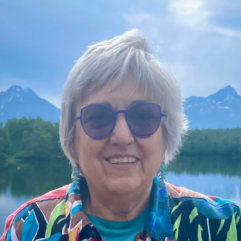 Smiling woman with short gray hair and blue sunglasses, wearing a colorful shirt, outdoors by a lake with mountains in the background.