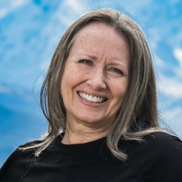Smiling woman with shoulder-length gray hair wearing a black top, against a bright blue sky.