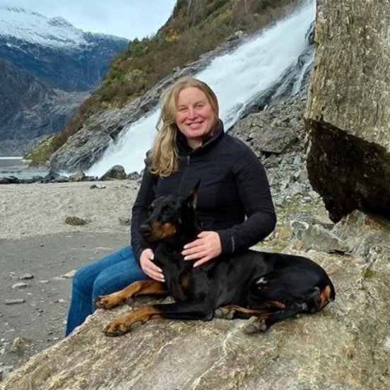 Woman sits on a rocky shore with a black and tan dog resting beside her, a waterfall behind them.