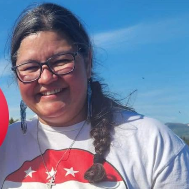 Smiling woman with glasses outdoors on a sunny day, wearing a white shirt with a red graphic and a braided ponytail.