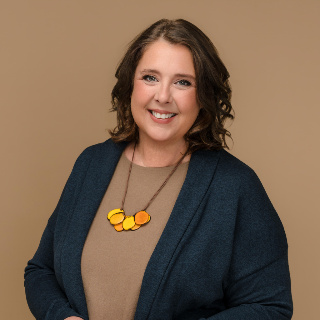 Smiling woman with dark hair wearing a blue blazer and tan top, accented by a yellow beaded necklace against a tan background.