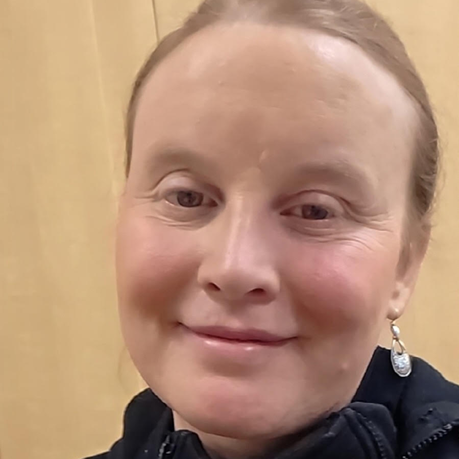 Close-up portrait of a smiling woman with light skin and earrings, wearing a dark jacket indoors.
