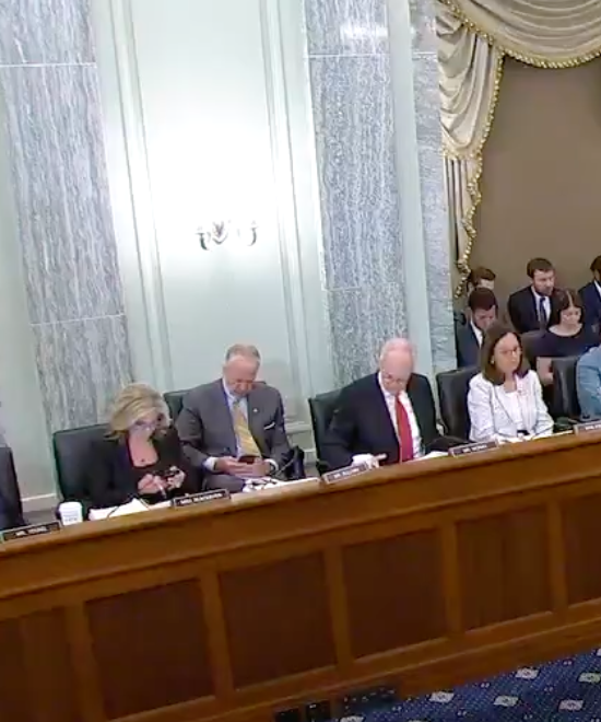 Panel of officials seated at a long wooden table in a formal hearing room, with documents and nameplates in front of them.