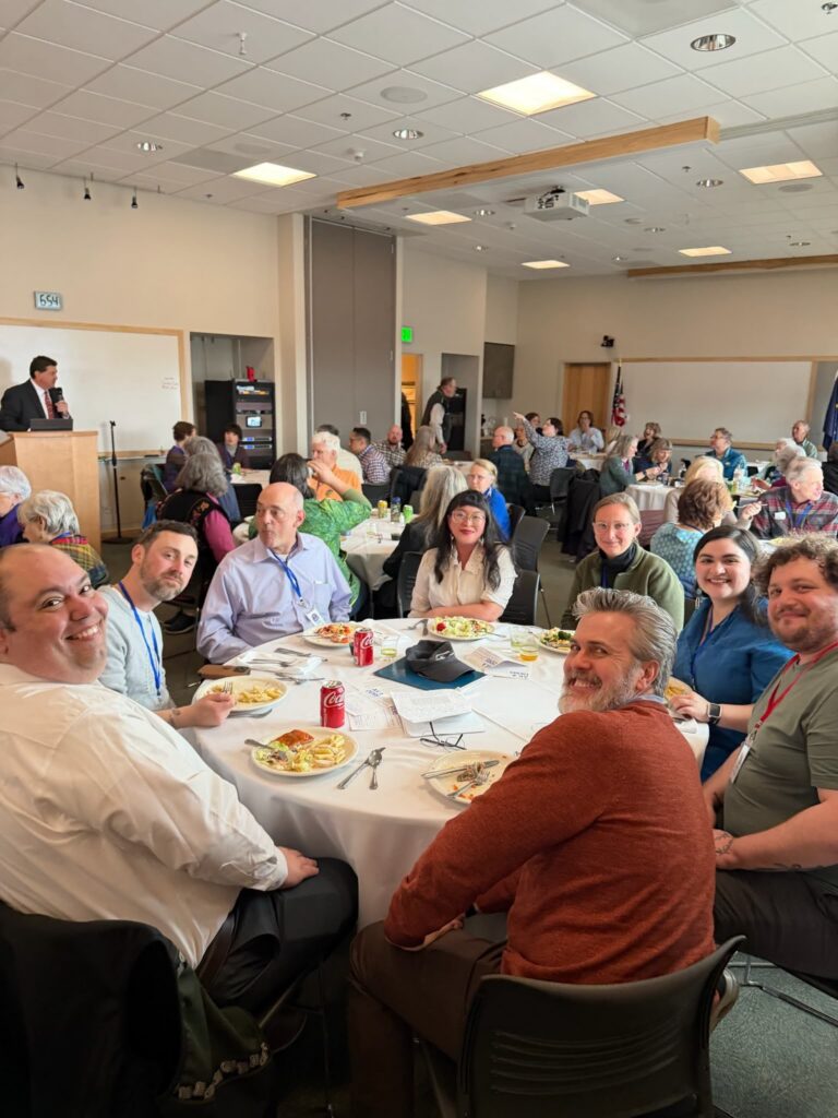 Group of people seated at round tables in a conference room, with a speaker at a podium on the left and attendees eating.