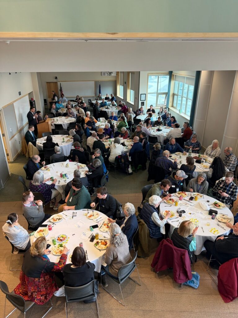 Large group of people dining at round tables in a bright banquet hall set for a luncheon