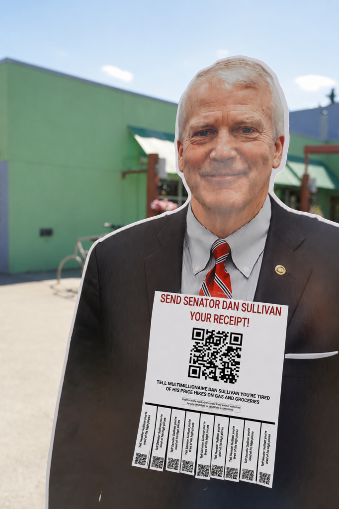 Cardboard cutout of a man in a suit and red striped tie, holding a flyer with a large QR code and the message 'Send Senator Dan Sullivan Your Receipt!'