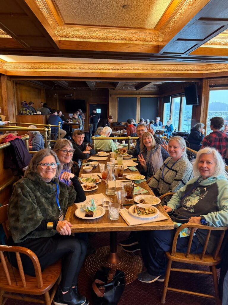 Smiling diners at a long wooden table in a warmly lit, wood-paneled restaurant with ornate ceilings and lakefront windows behind them.