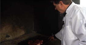 Chef in a white coat standing beside an open grill, inspecting slabs of meat in a dark barbecue pit.
