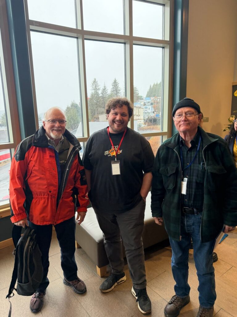 Three men pose indoors by a large window; left man in a red jacket with a bag, middle man smiles with a lanyard, right man in a black beanie and glasses with badge.