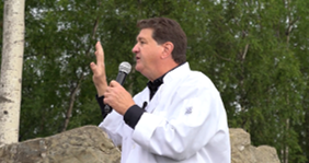 Man in a white shirt speaks into a handheld microphone outdoors, gesturing with his left hand.