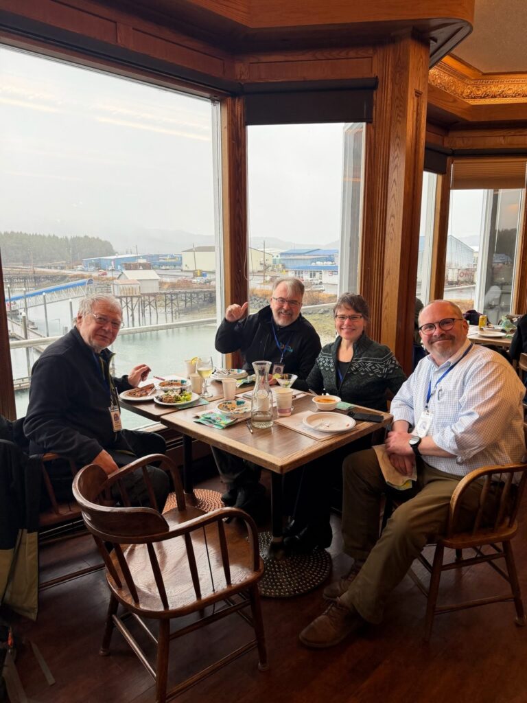 Four adults sit around a wooden table in a waterfront restaurant, smiling at the camera with plates of food and drinks in front of them while large windows overlook the harbor.