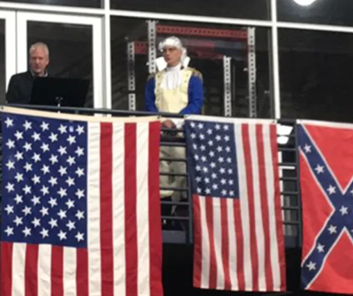 Two men stand on a balcony behind a row of American flags; one in a white wig and blue-and-gold costume, the other at a laptop, with a Confederate flag visible on the right.