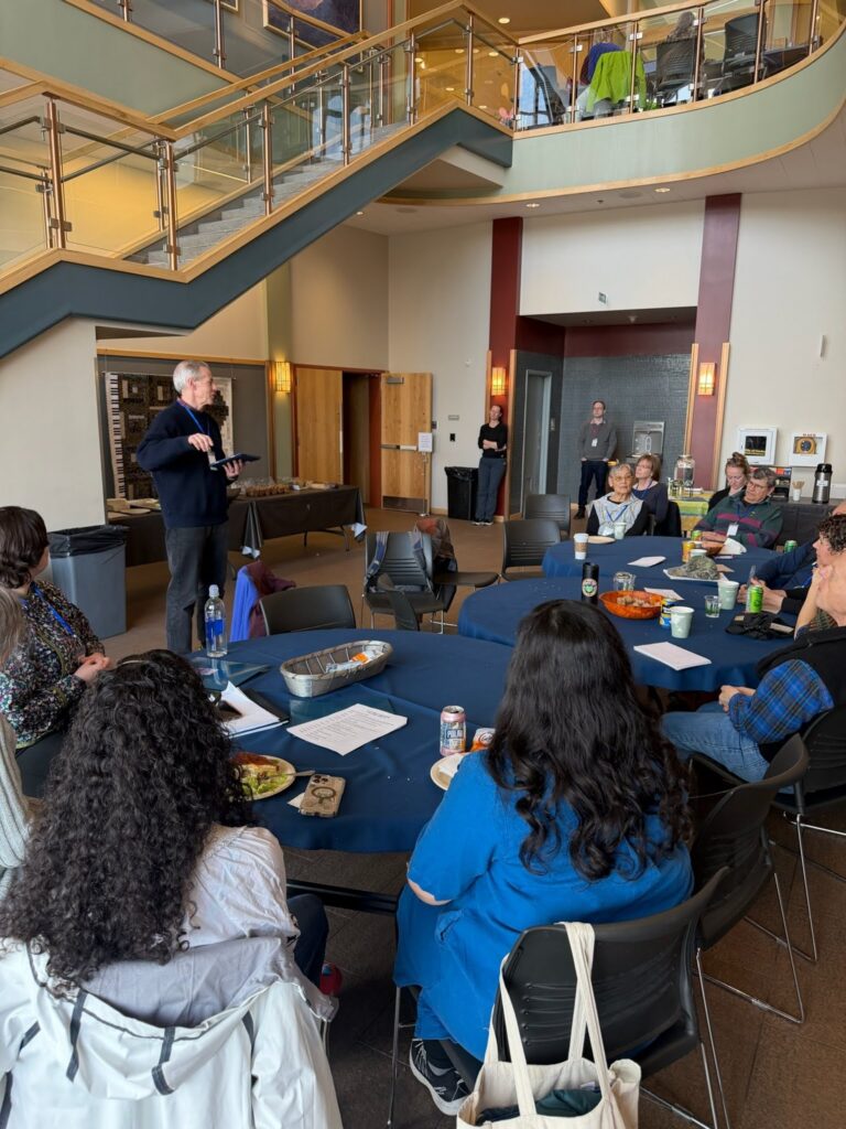Speaker standing at front of a room with round tables and attendees, giving a presentation in a lobby-like space with a curved glass staircase overhead.