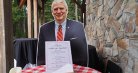Older man in a dark suit and red tie sits at an outdoor dining table with a sign/menu on the table.