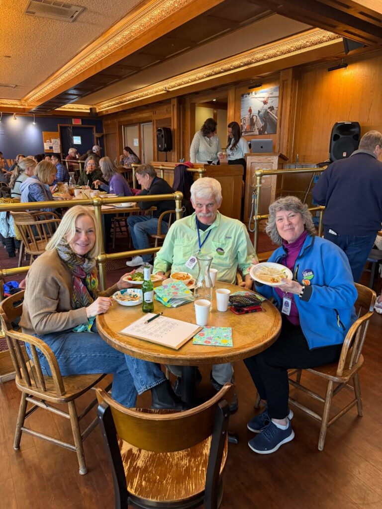 Diners enjoying a meal in a warm, wood-paneled restaurant; three people seated at a foreground table with plates and drinks, while others sit at tables in the background.