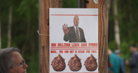 Poster on a wooden pole at an outdoor event showing a man with his hand raised and three pinecone illustrations beneath; crowd in background.