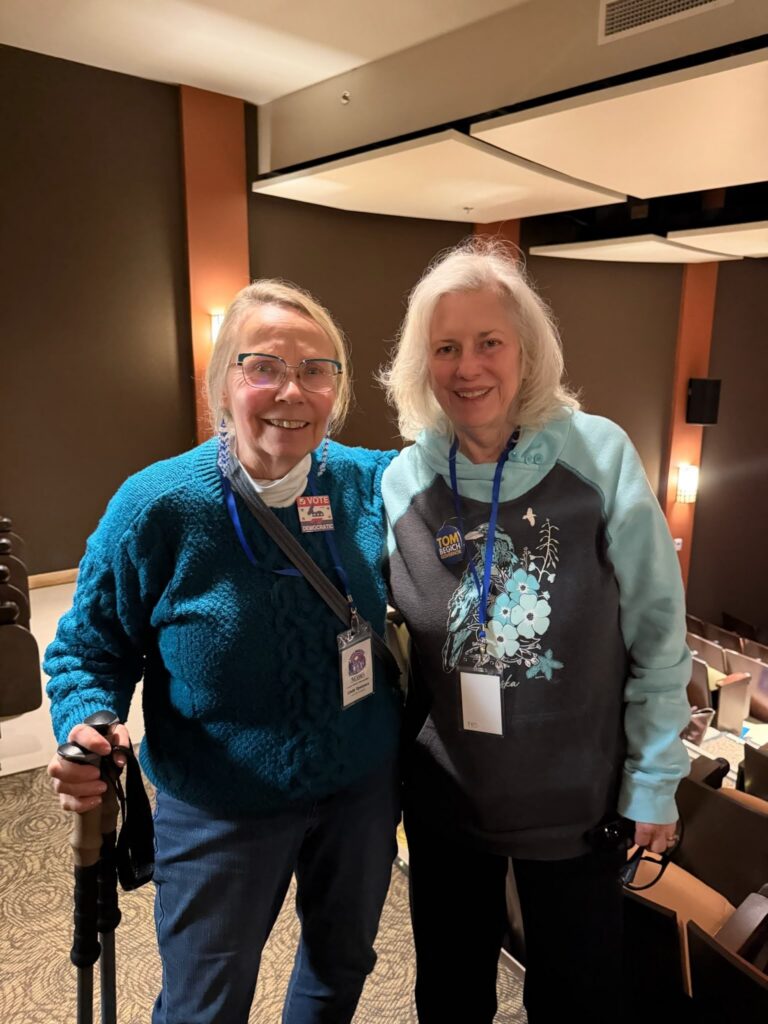 Two elderly women with conference badges smile for a photo in a meeting room; one holds a cane.