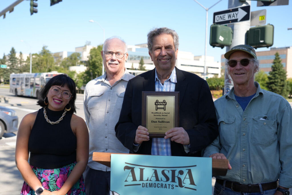 Smiling group of four outdoors; man in suit holds a plaque, Alaska Democrats sign visible in front.