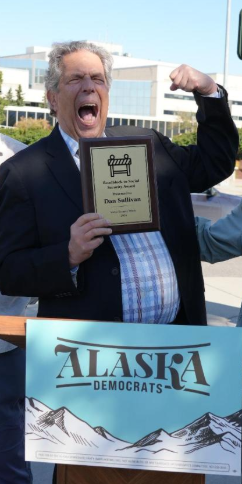 Man in a suit raises a clenched fist and shouts while holding a plaque, standing behind an Alaska Democrats banner with mountains.