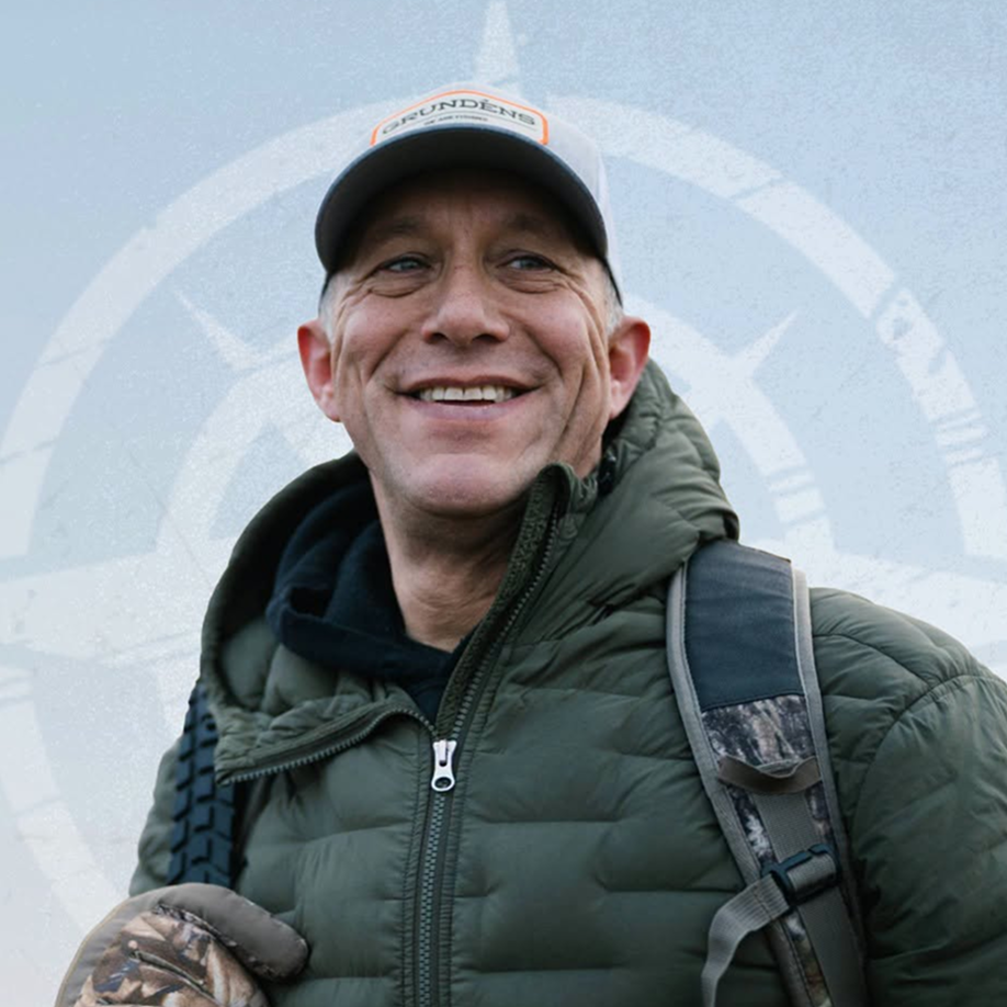 Smiling man wearing a gray cap and green puffer jacket with a backpack outdoors.