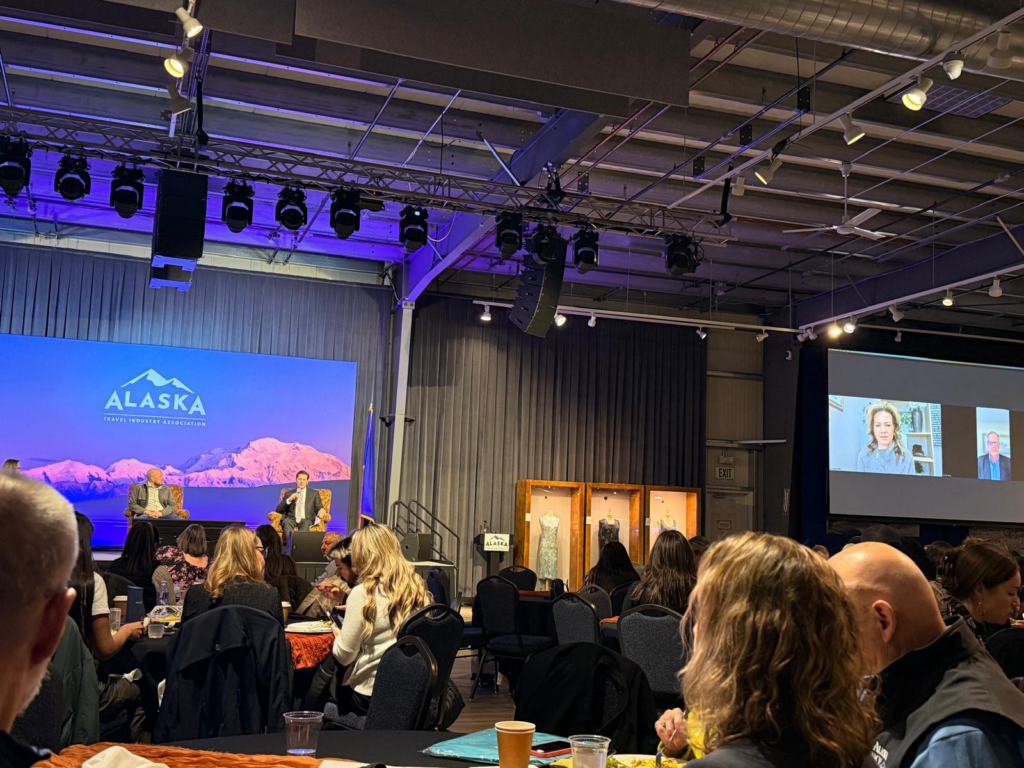 Stage at Alaska Travel Industry Association event with two speakers seated on chairs, blue backdrop, Alaska logo, and an audience in front.